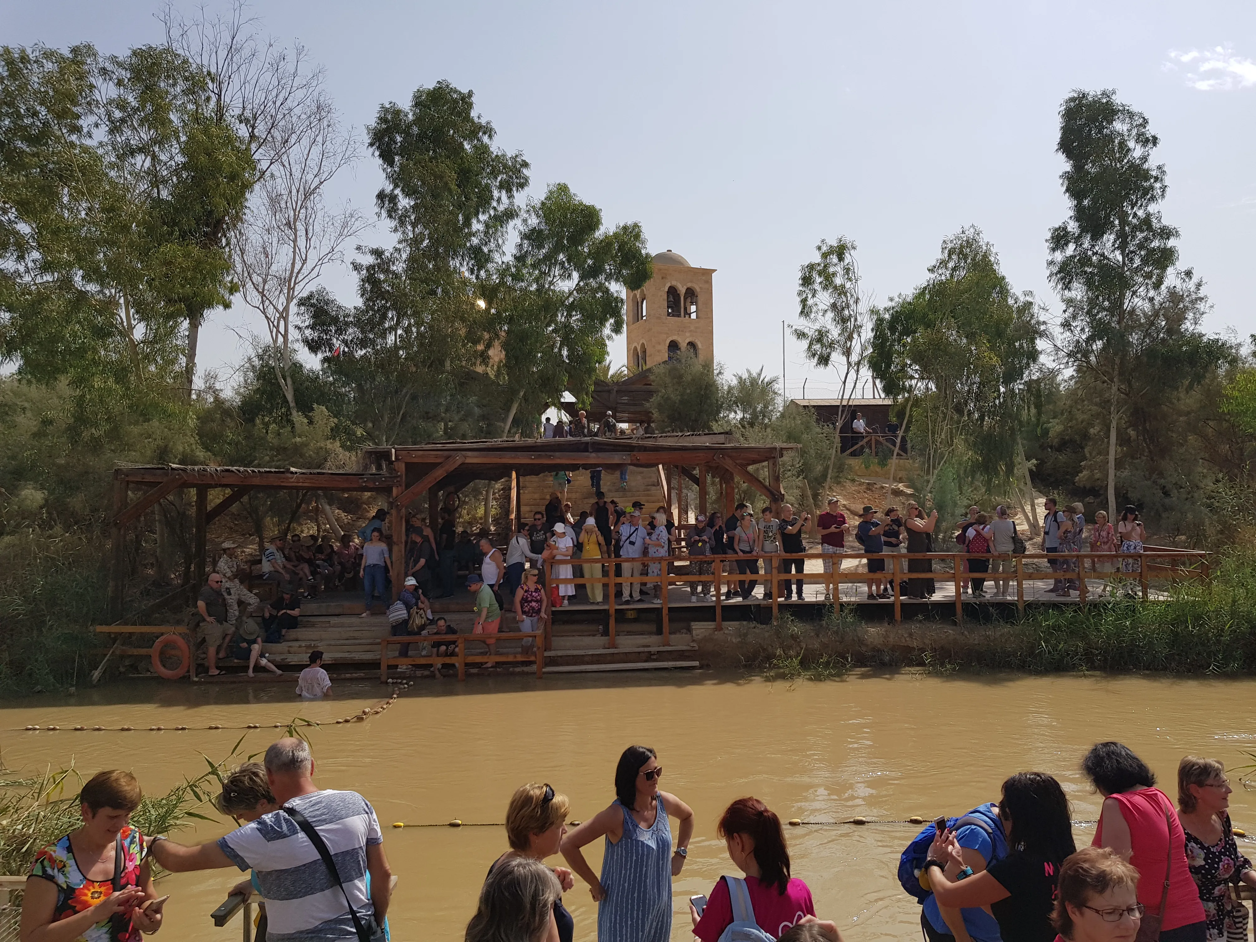 Peregrinos con túnicas blancas entrando al río Jordán en el sitio de bautismo de Qasr el-Yahud, con la orilla jordana visible al otro lado del agua