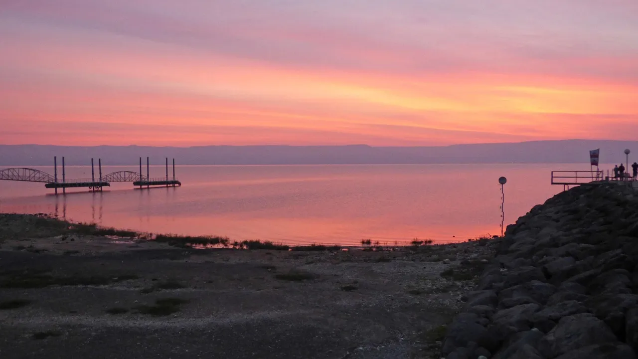 Mar de Galilea al amanecer, con el agua tranquila reflejando la luz dorada de la mañana y las alturas del Golán al fondo