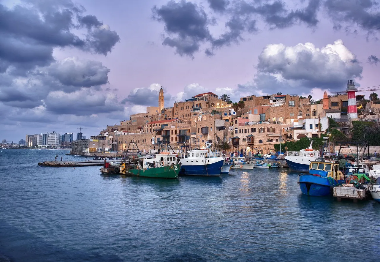 The ancient port of Jaffa at sunset, with stone quays, moored fishing boats, and the Ottoman-era clock tower visible against a warm sky